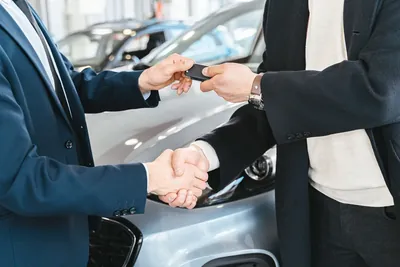 A car key being handed over during a handshake between two people outside a dealership