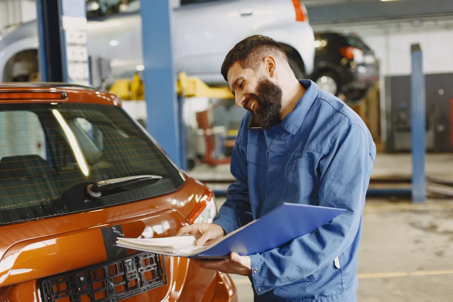 A mechanic in blue overalls holding a service folder beside a car in a workshop