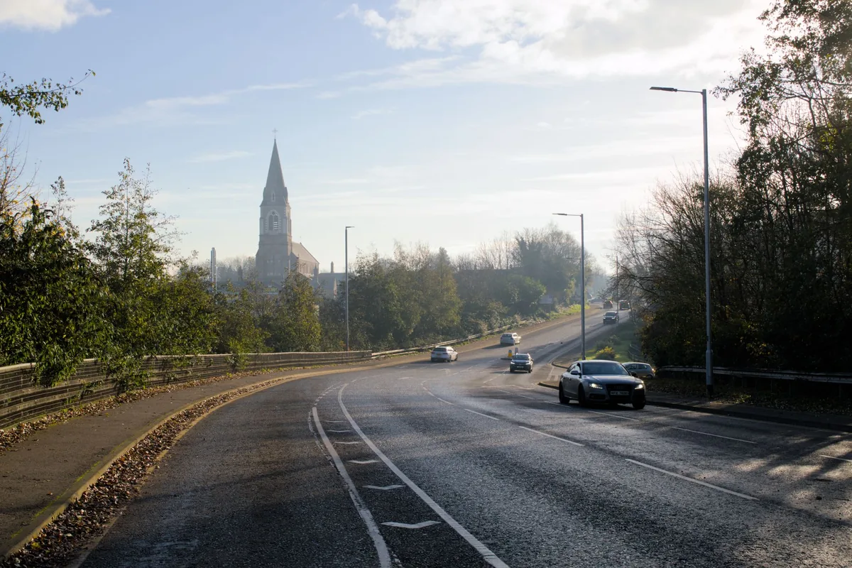 A car driving on a damp autumn road past a church spire in an Irish town