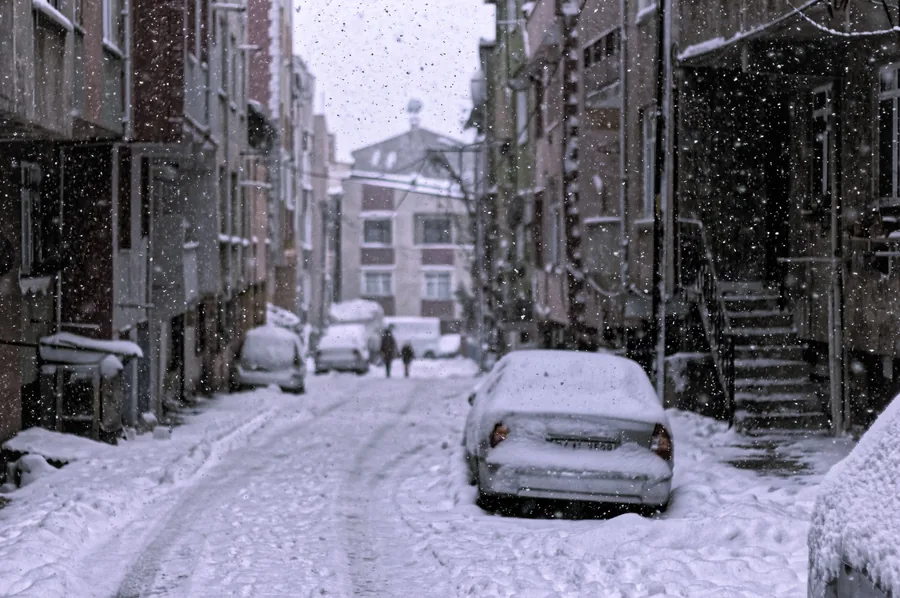 Snow falling on a narrow city street with cars parked along both sides under fresh snow