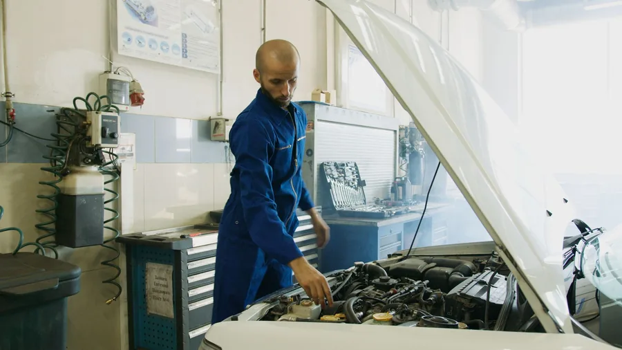 A mechanic in blue overalls inspecting a car engine bay in a workshop