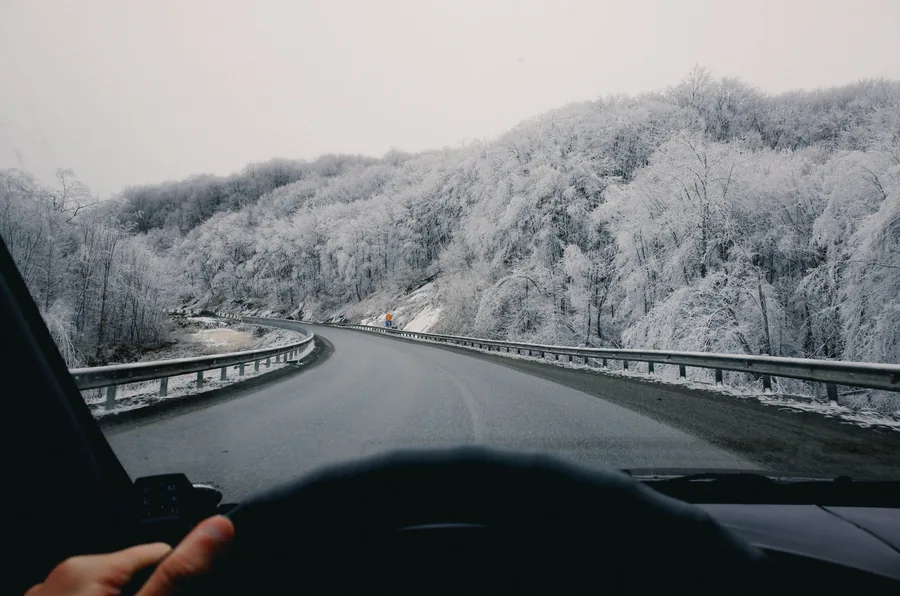 View through a windscreen down a wet winter road lined with snow-covered forest