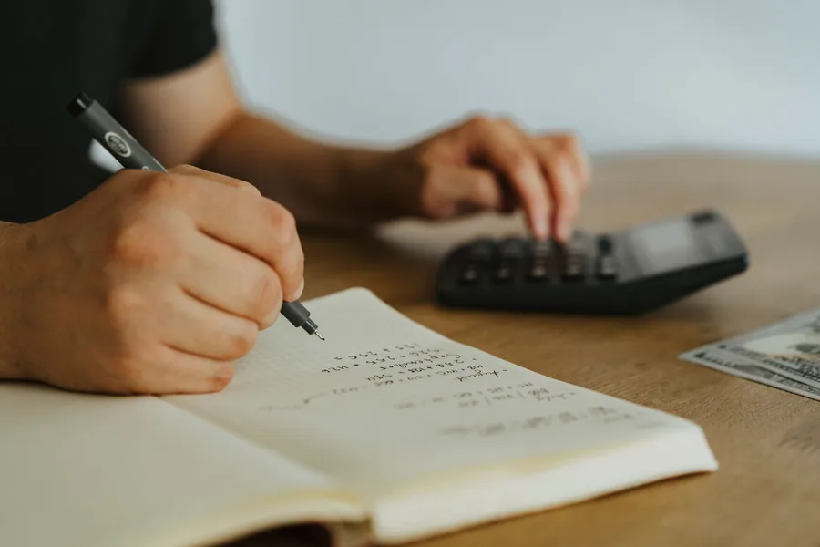 A person writing calculations in a notebook beside a calculator