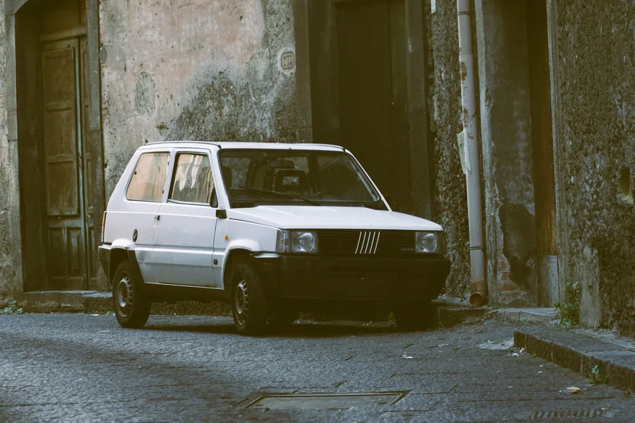 A small white city hatchback parked in a narrow paved alley between stone walls