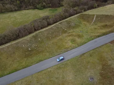 A single car driving along a narrow country lane between hedgerows and open fields